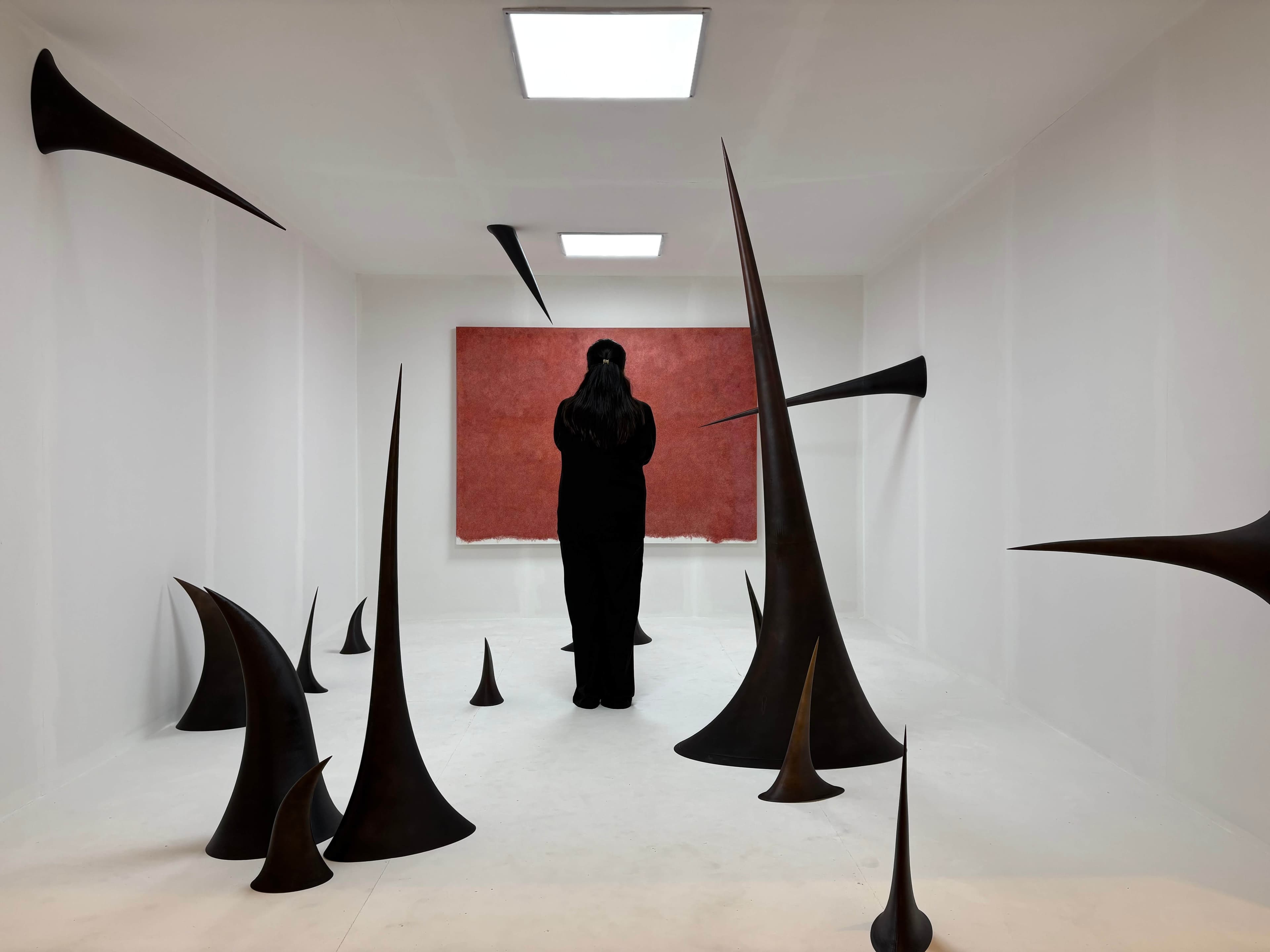 A figure in black faces a large red canvas in a white gallery, surrounded by tapering dark forms extending from the floor, walls and ceiling.